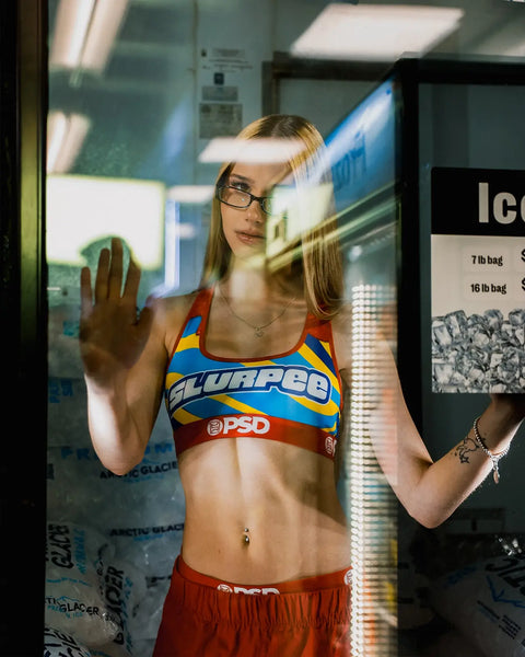 Woman wearing Slurpee-branded sports bra and shorts standing in front of refrigerated display case.